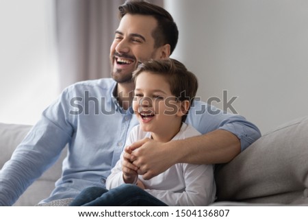 Happy young dad and cute small kid son laughing watching funny comedy movie tv show sitting on sofa, cheerful father having fun with child boy viewing television together at home relaxing on couch