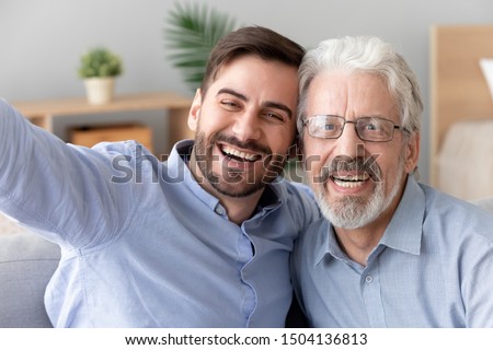 Happy two generations men old father laughing embracing young son take selfie shoot vlog together on phone looking at camera, senior dad hug adult man having fun making self portrait, mobile cam view