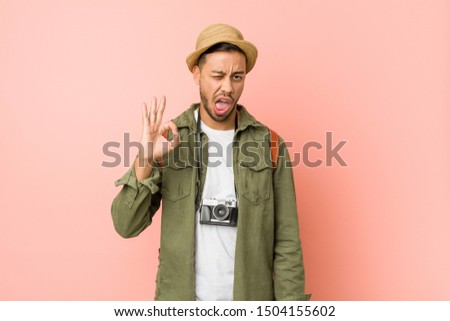 Young filipino traveler man winks an eye and holds an okay gesture with hand.