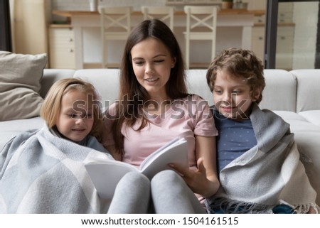 Young positive mommy sitting on warm heated floor with covered in plaid little adorable smiling kids siblings, reading fairy tale together. Happy mother holding book, telling story to cute children.