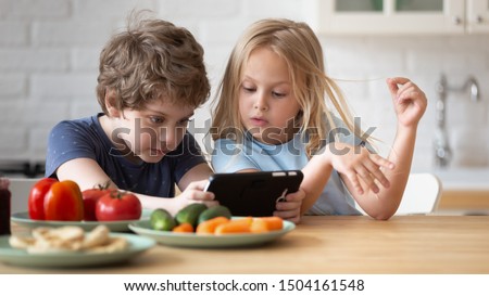 Close up head shot little adorable brother and sister using smartphone, playing, watching cartoons together. Involved in mobile game addicted to technology small children sitting at table on kitchen.