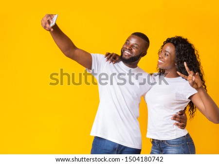 African couple in love making selfie over yellow studio background, girl showing victory sign, copy space
