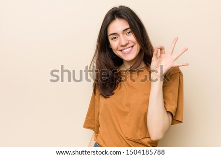 Young brunette woman against a beige background winks an eye and holds an okay gesture with hand.
