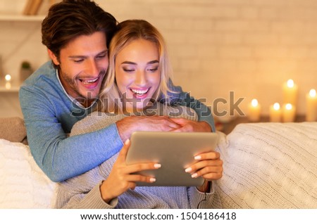 Happy Couple Using Digital Tablet Watching Movie Sitting On Couch At Home In Evening. Free Space