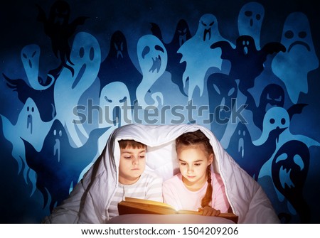 Enthusiastic little girl and boy reading book in bed after bedtime. Kids in pajamas with flashlight hiding under blanket together. Fearful children and imaginary monsters on deep blue background.