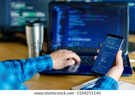 Closeup freelancer hand is holding smartphone with program code. Programmer developer is writing software on multiple computer screens at home office. Devices for working. Geek workplace concept. Royalty-Free Stock Photo #1504251140