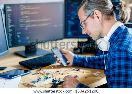 Developer is connecting breadboard to microcontroller. Man is holding smartphone with program code software for controlling electronic device. Chips, resistors, diodes on desktop of hardware engineer. Royalty-Free Stock Photo #1504251200
