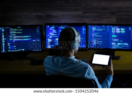 Software developer freelancer with white headphones and glasses working with program code: C++, Java, Javascript on wide displays and tablet at night. Develops new web, desktop, mobile application.