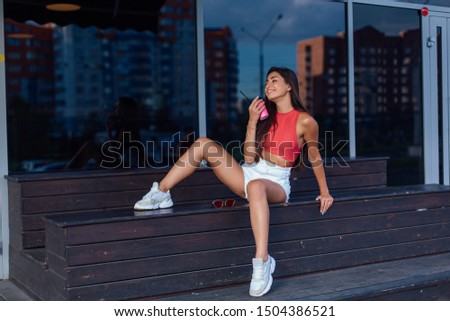 Stylish happy young woman wearing white shorts and sneakers holding pink cup of coffee to go sitting next to coffee shop. Portrait of smiling girl sitting on the bench with coffee.