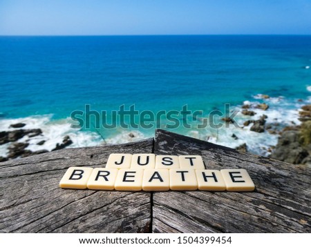 Letter tiles form the words "JUST BREATHE" on a wooden plank background, high on a cliff with the bright blue ocean, rocks, trees and grasses as backdrop, for a sign, banner, wallpaper or image 