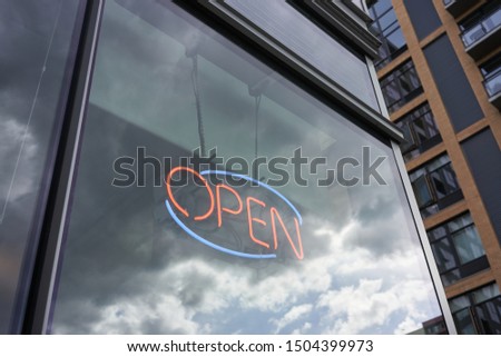 Blue and red lit open neon sign in a shop window against reflections of dark clouds and a gradually clearing sky. Open for business in a difficult time with determination and hopefulness.