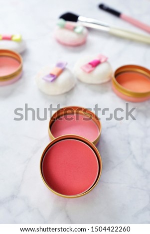 Small round blusher pots with different shades of pink and cute powder puffs with ribbons and makeup brushes in background