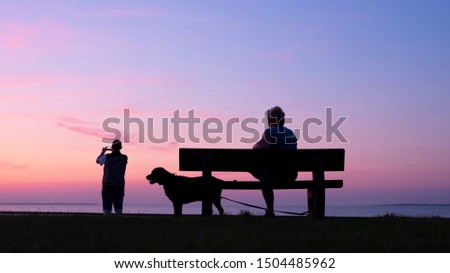 man takes pictures with moblie phone of colorful sunset while woman sits with dog on bench