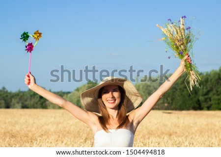 Young beautiful woman in a white dress and hat holds a bouquet with wildflowers and a toy visor on a milf's field. Concept of outdoor recreation, a trip to the village