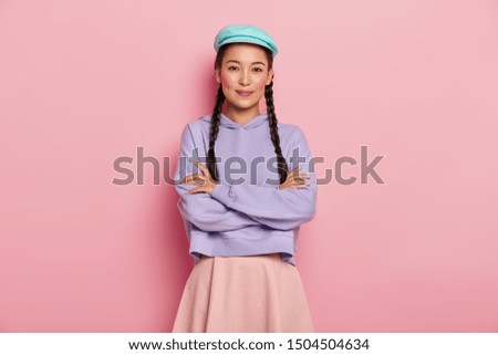 Pretty young female model with satisfied expression, keeps arms folded over chest, looks positively at camera, wears stylish cap, jumper and skirt, has two pigtails, isolated over pink studio wall