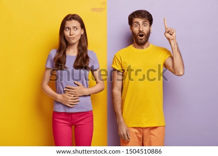 Hungry dark haired woman touches stomach, wants to eat something tasty, wears purple t shirt and pink trousers, impressed unshaven man in casual wear points index finger above, shows direction up Royalty-Free Stock Photo #1504510886