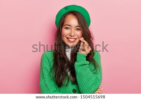 Charming young woman with dark long hair makes korean love sign, shapes heart with fingers, wears bright fashionable green beret and jumper on buttons, isolated over pink background. Hand gesture