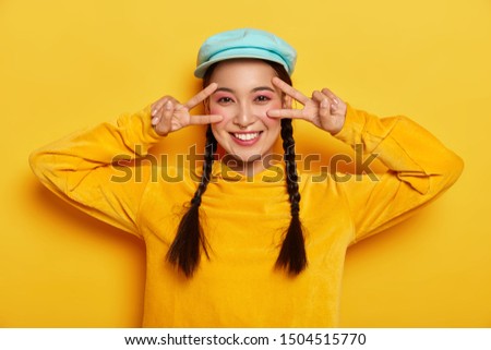 Horizontal shot of carefree smiling Asian lady makes victory peace gesture near eyes, has happy mood, smiles gently, wears vivid makeup, wears stylish hat and sweatshirt, isolated on yellow wall