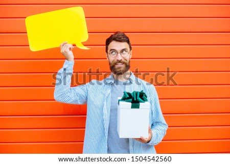 Attractive young beard man in cirlce glasses with think emotion on his face holding a speech bubble and present box with gift looking at camera and smiling, on red background outdoor portrait.