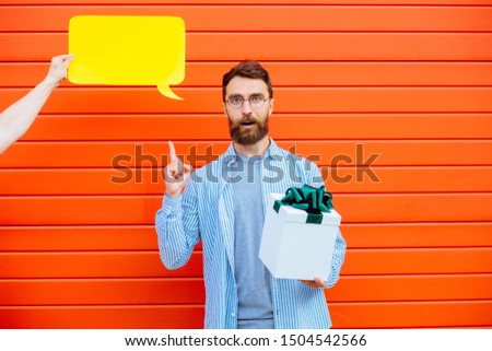 Hand of an unrecognizable person holding yellow speech bubble near head of beard man with different emotions on his face, with present box on red background. Make you think, manipulation concept.
