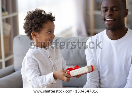 Little son making surprise for smiling African American father with closed eyes, preschool boy holding box, preparing to give gift to parent, congratulate with birthday or fathers day close up