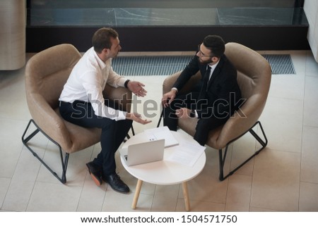 Top view two businessmen european and arabian ethnicity talking seated on armchair in modern office lobby, laptop relevant documents on coffee table, discuss plan future cooperation at meeting concept Royalty-Free Stock Photo #1504571750
