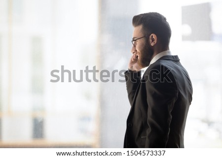 Bearded businessman middle eastern ethnicity wearing formal elegant black suit looks out window talk on phone, negotiating, communicates with corporate clients, having successful career growth concept