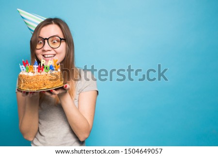 Crazy funny girl in a paper hat and glasses holding a big birthday cake on the blue background. Concept of prank and greetings.