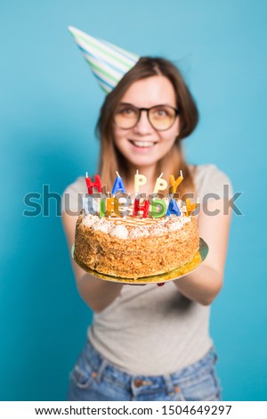 Close up funny positive girl in glasses and greeting paper hat holding a happy birthday cake in her hands standing on a blue background.