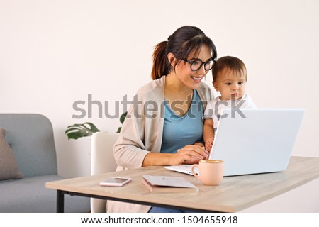 Stay at home mom working remotely on laptop while taking care of her baby. Young mother on maternity leave trying to freelance by the desk with toddler child. Close up, copy space, background.