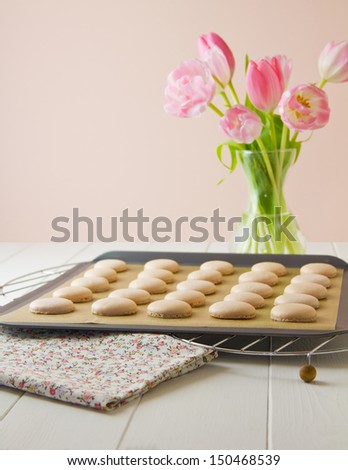 French macaron shells on baking sheet lined with baking parchment paper, on wire cooling racks. On white wood table with floral tea towel. Soft pink and white tulips sit to one side.