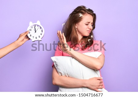 Studio portrait of dissatisfied young lady holds white pillow, waves aside alarm clock, unhappy to have not enough sleep, tired to wake up early, needs more rest, has feathers in her messy hair.