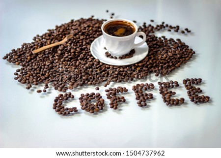 Letters made of beans and white cup.Coffee beans and white mug on bright background.White cup of black drink and brown beans. Black coffee in white cup and coffee beans text ''coffee". Space for text
