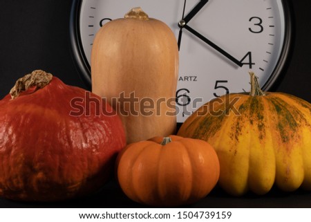 Cropped section of an arrangement with a white clock, red pumpkin, a mini orange pumpkin, butternut and a yellow pumpkin. Isolated on black background, close-up for concept.