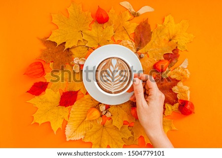 Autumn flat lay composition with dry leaves wreath frame and coffee latte cup in woman hand on bold orange color background. Creative autumn thanksgiving, fall, halloween concept. Top view, copy space