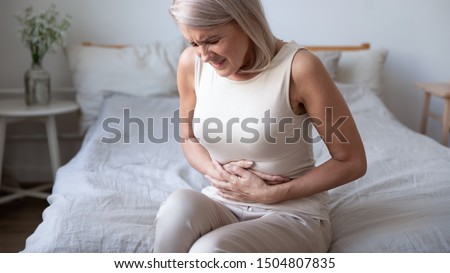 Unhealthy mature woman holding belly, feeling discomfort, health problem concept, unhappy older female sitting on bed, suffering from stomachache, food poisoning, gastritis, abdominal pain, climax Royalty-Free Stock Photo #1504807835