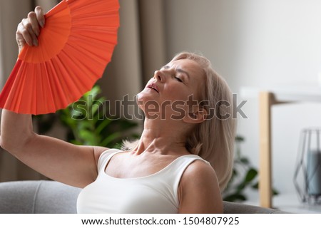 Exhausted older woman waving blue fan close up, suffering from heat, feeling unwell and uncomfortable, feeling uncomfortable, sweaty mature female cooling in hot summer weather, high temperature Royalty-Free Stock Photo #1504807925