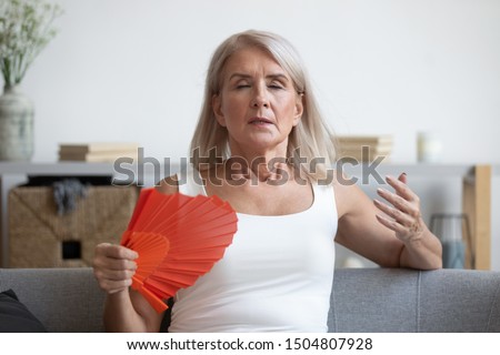 Tired mature woman suffering from heat at home, feeling uncomfortable, waving blue fan, sitting on couch, enjoying fresh air, sweaty older female cooling in hot summer weather, high temperature Royalty-Free Stock Photo #1504807928