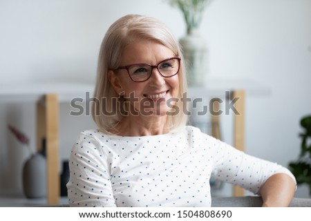 Head shot portrait close up smiling mature woman wearing glasses looking in distance, dreaming, thinking of future, older female with healthy smile sitting on couch at home, posing for photo