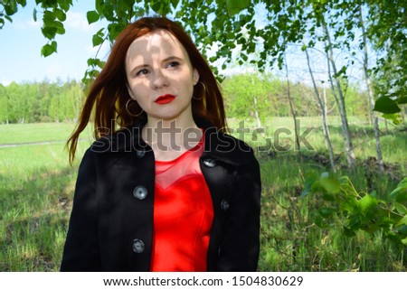 View through the green foliage of a red-haired girl in a coat in nature

