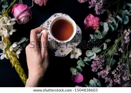 Woman's hand and with beautiful cup of tea on the table among colorful flowers, flat lay
