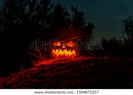 Carved halloween pumpkin jack-o-lantern wearing witch hat with burning candles glows in darkness. Spooky landscape
