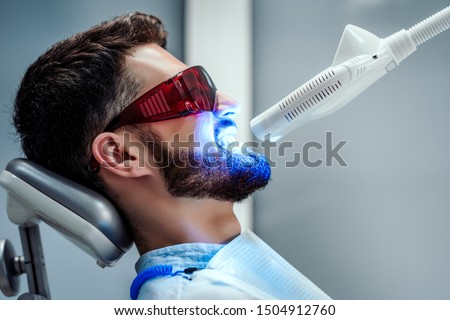 Unrecognizable dentist whitening teeth of young man sitting in dental chair in modern clinic. Side view. Royalty-Free Stock Photo #1504912760