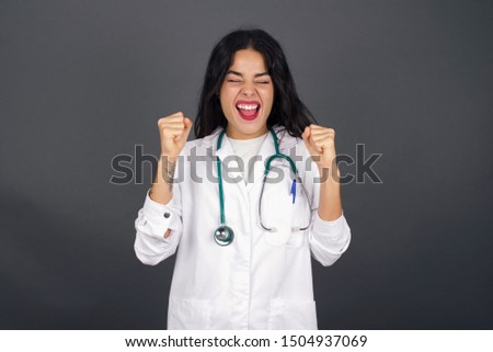 Caucasian brunette doctor woman rejoicing her success and victory clenching her fists with joy. Lucky woman being happy to achieve her aim and goals. Positive emotions, feelings.