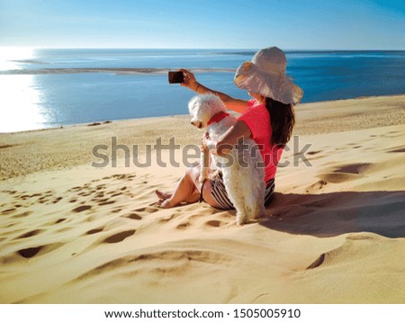 Beautiful woman using smart phone with sea view background. Sitting on sandy beach, next to her her little cute white dog.