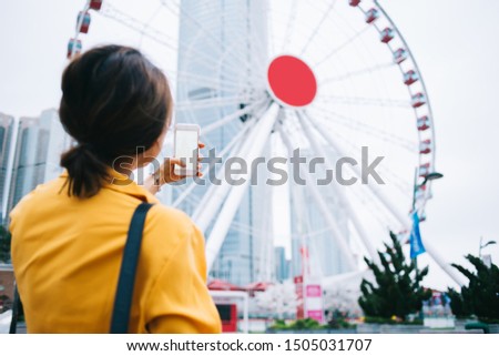 Back view of modern trendy woman in casual outfit holding smartphone with blank screen and picturing modern Ferris wheel on Hong Kong street park