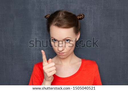 Studio close-up portrait of dissatisfied serious woman wearing orange T-shirt, with funny hairstyle, shaking finger while making reprimand or rebuke, looking strictly at camera, over gray background