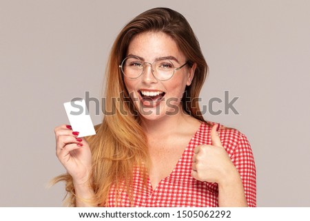 Beautiful young redhead woman in casual clothes holding credit bank card in hand, smiling to the camera, posing in studio. People lifestyle concept. 
