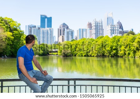 Young man sitting on railing in Piedmont Park in Atlanta, Georgia looking at cityscape skyline urban view with city skyscrapers downtown by Lake Clara Meer