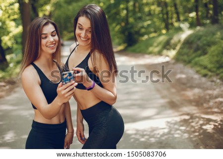Two girls standing in a summer forest. Friends have fun in a park. Sports ladies in a black sportsuit. Women with phone
